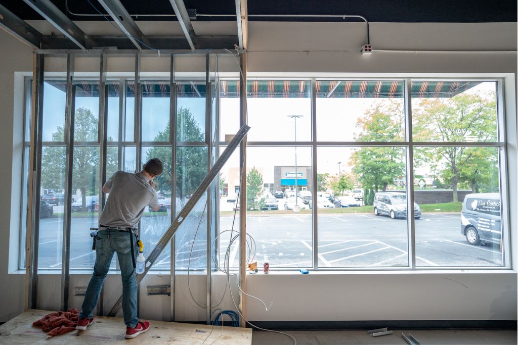 A worker installs window film on large commercial windows inside an office under construction, with tools and building materials nearby. The film is being applied to improve glare reduction and energy efficiency.