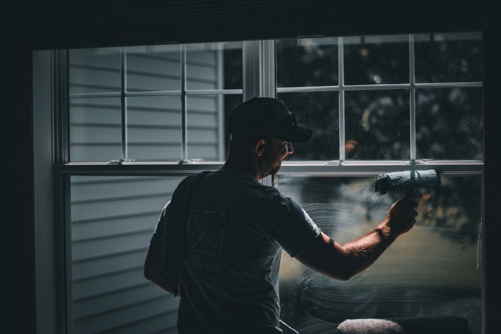 A worker applies window film to a residential window using a roller, with sunlight streaming through the glass. The process highlights residential window tinting aimed at improving energy efficiency and UV protection.