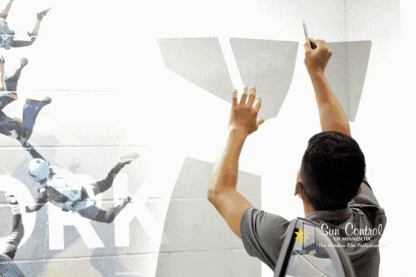 A Sun Control of Minnesota technician applies a large custom window graphic featuring skydivers and bold lettering to an interior glass wall. The technician uses a blade to trim the graphic film precisely along the edges.