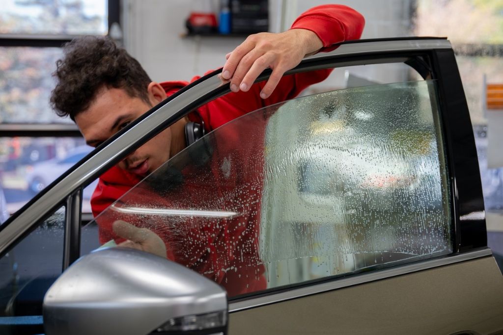 A technician in a red hoodie applies window tint film to a car door window, with water droplets visible on the glass surface during the installation process. The scene captures a careful, mid-application moment in a professional tint shop.